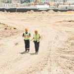 Two workers in safety gear walking across a construction site under a clear sky.