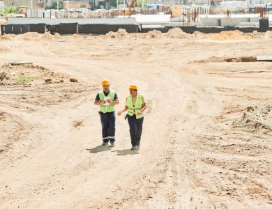 Two workers in safety gear walking across a construction site under a clear sky.