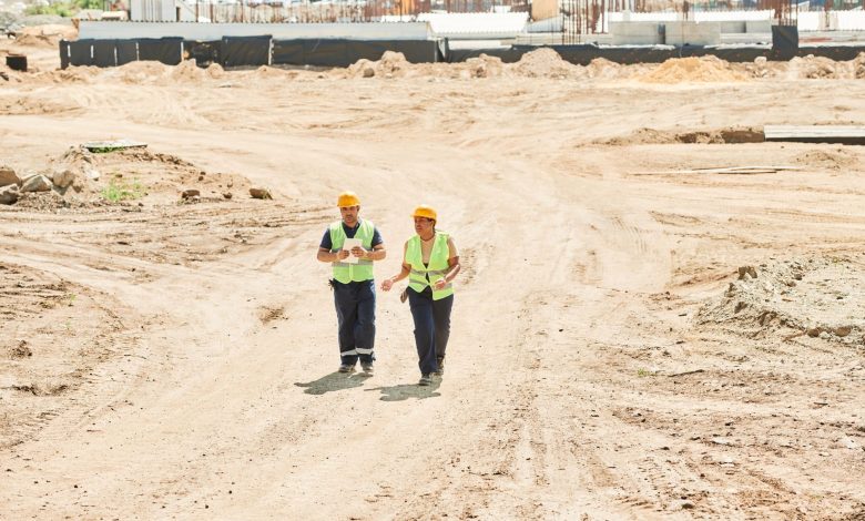 Two workers in safety gear walking across a construction site under a clear sky.